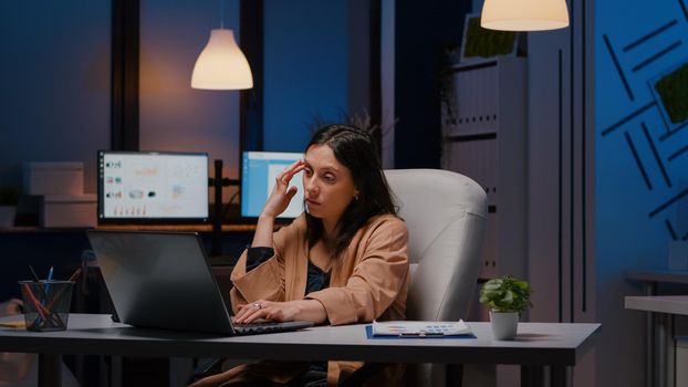 Overworked exhausted businesswoman working in startup office checking management strategy on laptop late at night. Stressed executive manager remains alone in company room after his collegue left