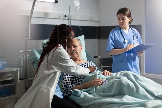 African doctor specialist using stethoscope listening senior man heart, laying in bed breathing with help from oxygen tube, and medical nurse is taking notes on clipboard.