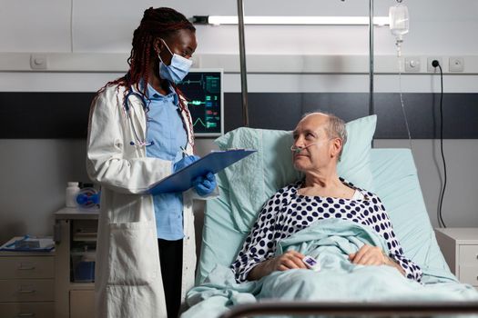 African american doctor with surgical mask reading diagnosis from clipboard to sick ill unwell senior patient laying in bed, breathing with help from oxygen mask, listening discussing with medical staff about recovery.