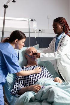 African doctor and medical assistant helping senior man breath using oxygen mask, in hospital laying in bed. Sick patient cant breath because of lung infection. Nurse listening heart with stethoscope.