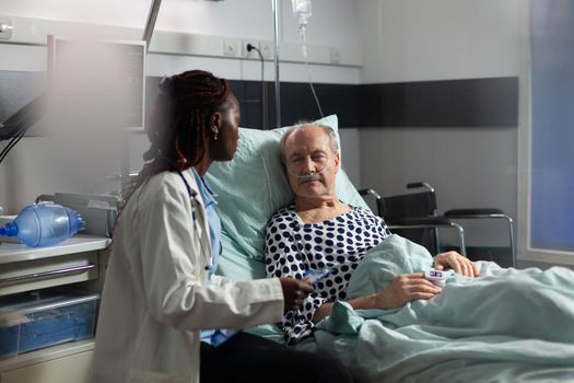 Unwell senior patient laying in bed breathing through oxygen test tube, listening african american doctor specialist, talking about treatment and recovery. Oxymeter attached on finger.