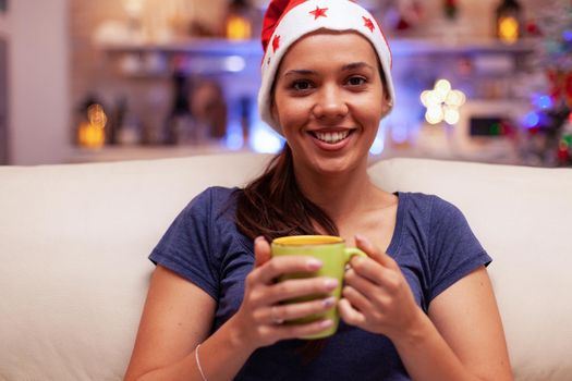 Portrait of smiling woman holding cup of coffee in hands during christmastime sitting on couch in xmas decorated kitchen. Adult person enjoying winter season celebrating christmas holiday