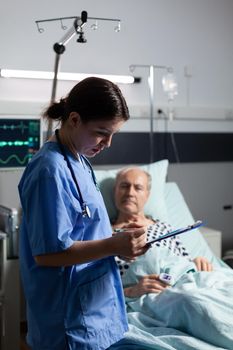 Medical nurse in scrubs taking notest on cliboard after consultation of sick unwell senior man. Patient with iv drip attached breathing with help from oxygen mask.