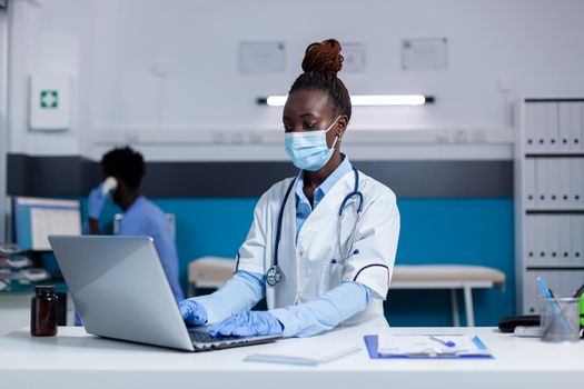 African american doctor sitting at desk with laptop computer in healthcare office. Black woman with medic profession wearing face mask and stethoscope for examination at medical clinic