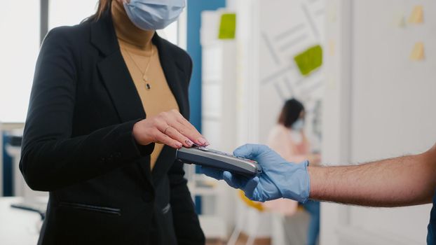 Closeup of businesswoman paying takeaway food order with credit card using POS contactless service during takeout lunchtime. Delivery man with medical face mask and gloves giving fresh food meal