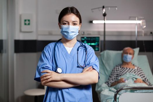 Portrait of medical nurse with chirurgical mask in hospital room with sick unweel senior pantient with trauma looking at camea with arms crossed, wearing scrubs.