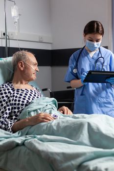 Medical assistant with protection mask checking treatment of senior man reading notes on tablet pc. Sick elderly laying in bed discussing with nurse about diagnosis expertise with iv bag attached.