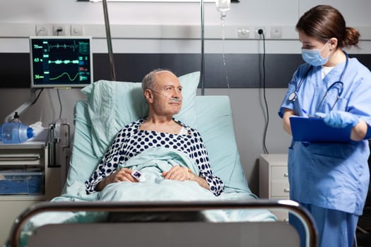 Medical nurse in scrubs with chirurgical mask taking notest on cliboard during consultation of sick unwell senior man. Patient with iv drip attached breathing with help from oxygen mask.