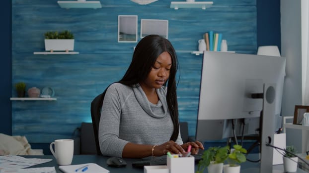 Black student sitting at desk table start working at online homework using computer typing information on keyboard searching webinar courses. African american young woman sitting at desk in living room