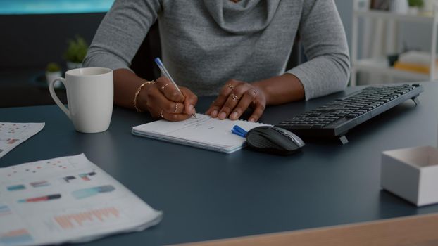 Closeup of student with black skin writing communication homework on notebook while sitting at desk in living room. Young woman studying math on elearning platform doing homework during high school