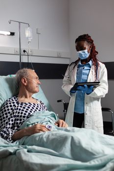 African american doctor with chirurgical mask in hospital room discussing diagnosis and treatment with sick senior man laying in bed reassuring full recovery. Unwell patient breathing through oxygen mask.