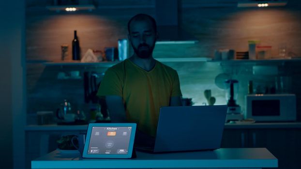 Man working from house with automation lighting system, controlling ambience with wifi gadget, sitting in kitchen turning on the lights. Person using voice command to smart home application on tablet.