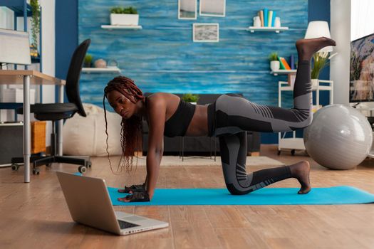 Black woman doing straight- leg donky kick pushing left foot up keeping leg bent, watching tutorial video on laptop screen, wearing sportwear on yoga mat for muscle strenght andn healthy lifestyle.