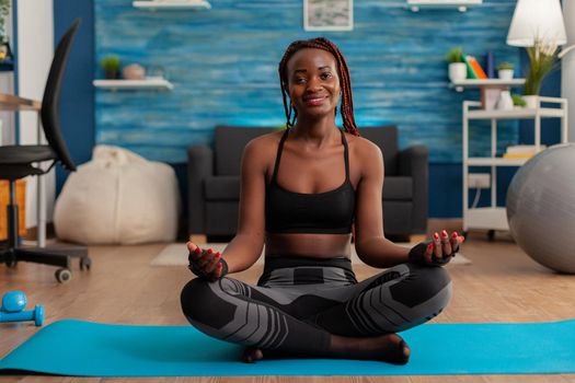 Portrait of black woman smiling practicing yoga at home sitting in lotus pose on mat mediting, practicing mindfulness meditation harmony, African american person doing relaxing workout.