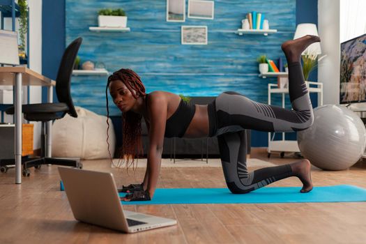 Black woman doing straight- leg donky kick pushing left foot up keeping leg bent, watching tutorial video on laptop screen, wearing sportwear on yoga mat for muscle strenght andn healthy lifestyle.