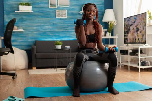 Joyful athletic black person in leggings using stability ball for training biceps, doing curls with dumbbells. Strong athletic person doing sports at home using modern equipment for