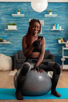 Sporty athletic black woman relaxing on stability ball checking social media using smartphone after intense workout exercising, in home living room sitting on fitness mat for muscle strenght.