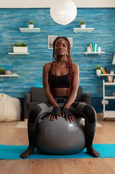 Sportive active strong black woman sitting on stability ball resting, after intense training in home living room. Slim fit african dressed in sportwear leggings.