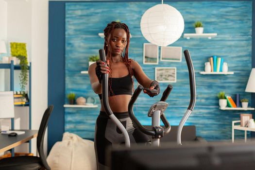 Strong athletic fit black woman doing cross training exercising in home living room using modern eliptic cardio equipment. exercising and changing tv channels.