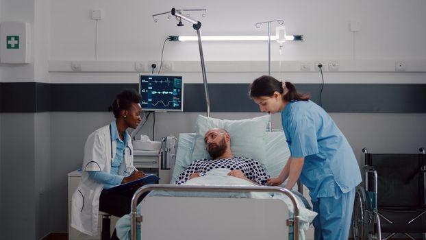 Practitioner afro american doctor discussing with sick man disease treatment monitoring symptom writing medical expertise on clipboard. Nurse arrange patient bed working in hospital ward