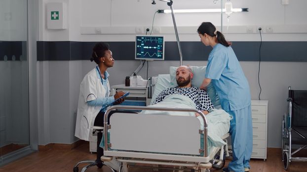 Afro american practitioner discussing sickness symptom with sick man writing medical expertise on clipboard working in hospital ward. Patient resting in bed while practitioner monitoring recovery