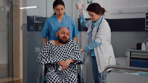 Sick man resting in bed during respiratory recovery in hospital ward during medical physiotherapy. Physical nurse helping patient putting in wheelchair after suffering leg fracture