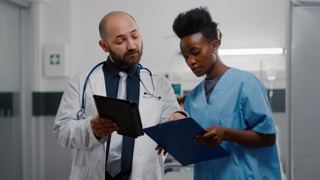 Practitioner doctor with afro american assistant analyzing recovery treatment using tablet computer working in hospital ward. Patient woman sitting in clinical bed discussing with specialist physician