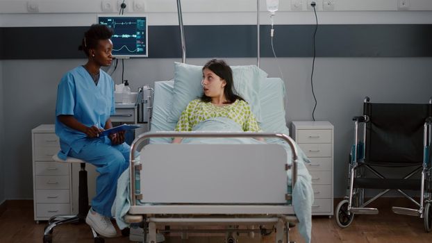 Sick woman adult resting in bed while black nurse analyzing disease expertise writing medical disease treatment on clipboard. Assistant discussing illness therapy working in hospital ward