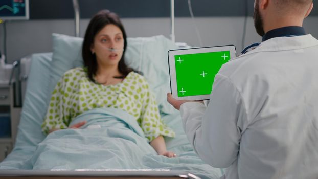 Physician practitioner man discussing with woman patient resting in bed about disease treatment. Doctor holding mock up green screen chroma key tablet computer with isolated display in hospital ward