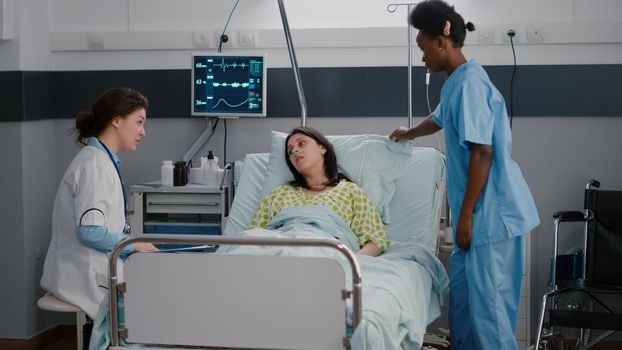 Nurse with dark skin helping patient to sit comfortable in hospital ward during disease recovery. Sick woman resting in bed while practitioner doctor discussing sickness treatment