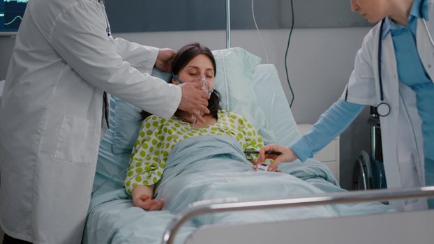 Medical team checking heartbeat of sick woman during respiratory healthcare working in hospital ward. Patient sitting in bed while doctor putting oxygen mask monitoring breath condition