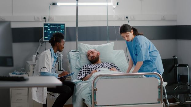 Patient talking with medical doctors while sitting in bed during disease recovery in hospital ward. Practitioner writing sickness treatment on clipboard checking heart pulse