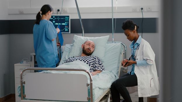 Specialist physician black woman doctor explaining medical diagnosis showing x-ray using tablet computer in hospital ward. Sick man patient sitting in bed during disease recovery treatment