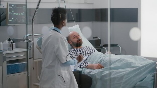 Afro american doctor checking sick man monitoring sickness symptom during recovery appointment in hospital ward. Patient sitting in bed while doctor analyzing heartbeat pulse