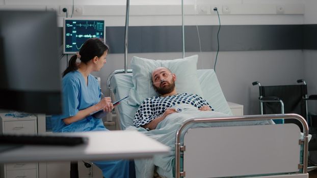 Sick man sitting in bed with oxygen tube explaining sickness symptom to medical nurse while writing disease treatment on clipboard. Medical nurse putting oximeter examining respiratory health