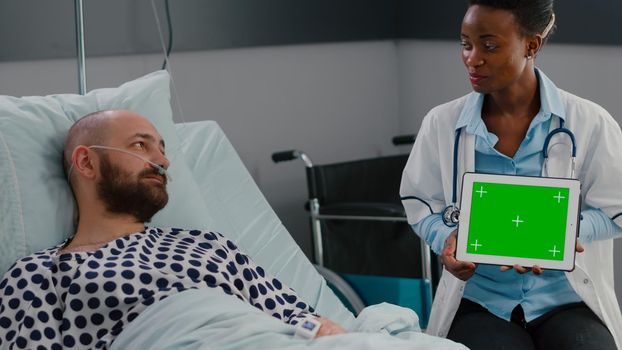 Afro american doctor in uniform discussing with sick man about sickness recovery treatment. Practitioner holding mock up green screen chroma key tablet computer with isolated display in hospital ward