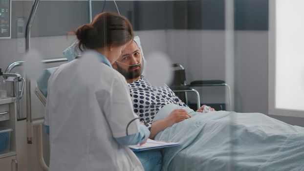 Professional doctors discussing sickness symptom with sick man writing medical tratment on clipboard working in hospital ward. Patient resting in bed while practitioner monitoring disease recovery