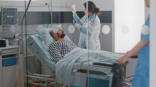 Medical team monitoring patient vital checking heart rate helping with fluids drip bag for respiratory therapy in hospital ward. Nurse putting in wheelchair for physiotherapy recovery