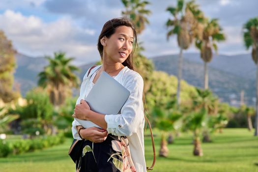 Outdoor portrait of young woman with laptop and backpack. Asian female adult student, creative freelancer, in tropical park, mountains background. E-learning, online technologies, education, business