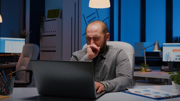 Exhausted sleppy businessman yawning while working at management project deadline late at night in start-up company office. Workaholic stressed entrepreneur typing financial strategy on laptop computer