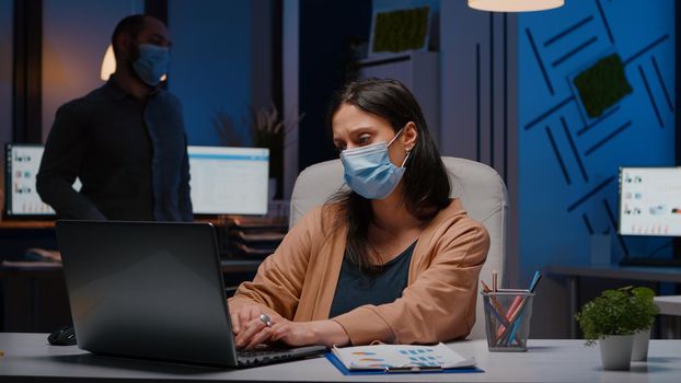 Workaholic businesswoman with face mask against covid19 working in startup office analyzing economic strategy late at night. Exhausted manager remains alone in company room after collegue left