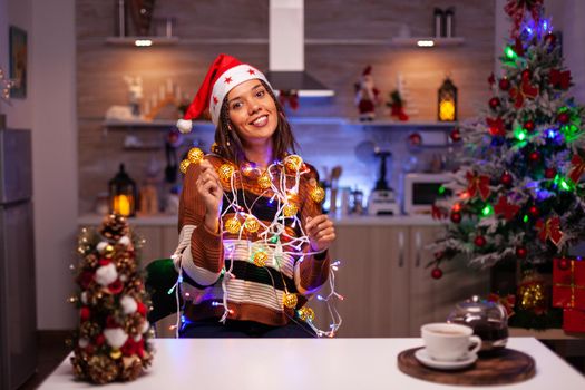 Young cheerful woman tangled in string lights used for christmas home decorating. Festive caucasian adult using wire garland with illuminated bulbs for kitchen decor and tree ornaments