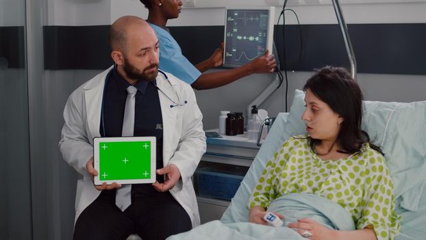 Specialist man in white coat talking with sick woman about illness recovery treatment. Practitioner holding mock up green screen chroma key tablet computer with isolated display in hospital ward