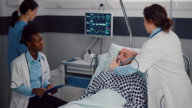 Afro american doctor discussing with sick man writing symptom sickness on clipboard while woman medic putting oxygen mask monitoring breath illness. Patient resting in bed in hospital ward