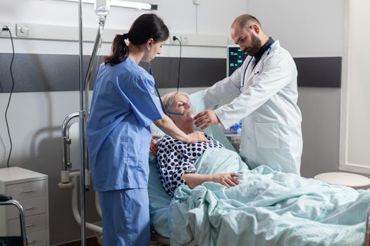 Unconscious senior woman patient laying in hospital bed and medical staff is helping her breath using oxygen mask. Doctor nurse using stethoscope listening heart of hospitalized elderly woman.