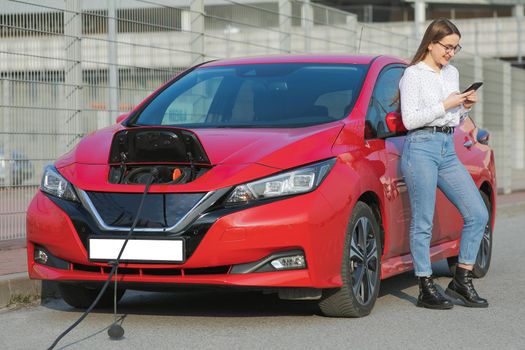 Connecting the Charger Plug of an Electric Car. Girl stands with phone near her electric car and waits when vehicle will charged.