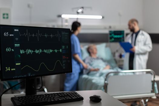 Monitor in hospital ward showing bmp from patient, laying in bed with oxymeter attached on finger, discussing with doctor and nurse about recovery treatment.