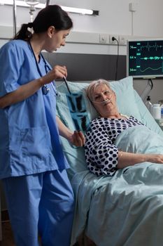 Medical nurse analyzing senior patient chest x-ray in hospital room, discussing explaining diagnosis. Old woman breathing through respiratory tube laying in bed listening health care specialist.