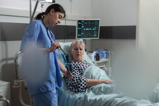 Medical nurse explaining chest x-ray radiography to elderly patient, breathing with help from oxygen mask because of respiratory illness. Getting medicine through iv drip bag.