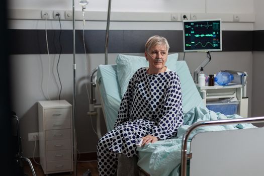 Senior woman patient sitting on the edge of bed in hospital bed looking pensive, breathing inhale exhale with help from oxygen mask, receiving medication through an intravenous line